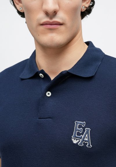 Πόλο μπλε ναυτικού χρώματος από υφασμάτινη υφή. Διαθέτει πατιλέτα με δύο κουμπιά και κεντητό λογότυπο "EA". Κλασικός σχεδιασμός για το γιακά.