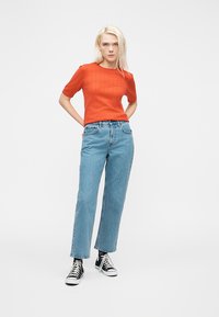 Νέα γυναίκα με ξανθά μαλλιά που φοράει πορτοκαλί κοντομάνικο μπλουζάκι, ανοιχτόχρωμο μπλε τζιν, μαύρες κάλτσες και μαύρα ψηλά αθλητικά παπούτσια, στέκεται με τα χέρια πίσω από την πλάτη.