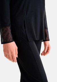 Zwarte tuniek met doorzichtige stippenaccenten op de mouwen en zijsplitten, gecombineerd met zwarte strakke broek. Fluweelzachte textuur overal.