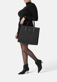 Zwarte tote bag van textuur stof, vierkante vorm en dubbele leren handgrepen. Beschikt over een subtiele logo aan de voorkant.