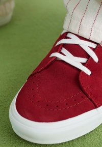 Røde ruskinds sneakers med hvide snørebånd, perforeringer på tåhætten og hvid gummisål. Sat op mod en grøn baggrund.