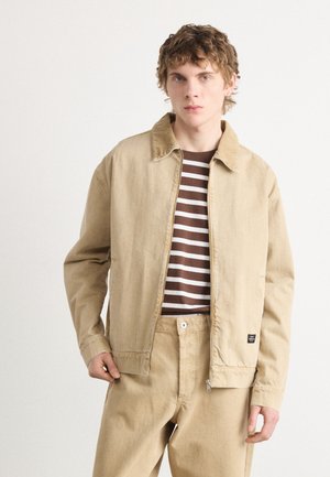 Giacca di cotone beige con chiusura a zip, colletto a punta e tasche laterali, indossata sopra una camicia a righe marroni e bianche e pantaloni beige coordinati.
