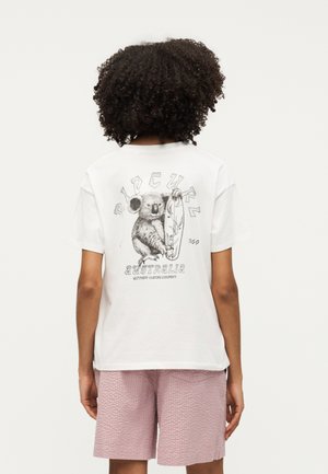 Άτομο με σγουρά μαλλιά που φοράει λευκό μπλουζάκι με έναν κοάλα που κρατάει σανίδα σερφ και το κείμενο "Rip Curl Australia Ultimate Surfing Company."