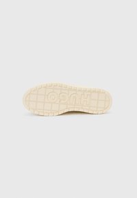 Suela de una zapatilla en goma beige claro con superficie texturizada; presenta el nombre de la marca "HUGO" en relieve de forma prominente.
