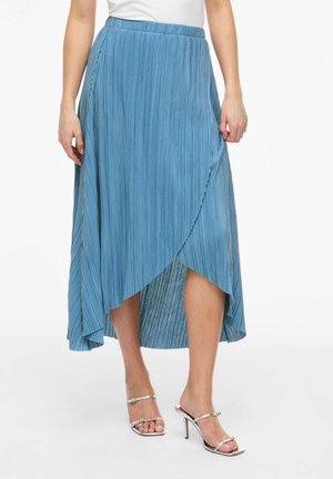 Nő kék pliszírozott, átkapcsolós midi szoknyában és ezüst magas sarkú szandálban, fehér háttér előtt állva.