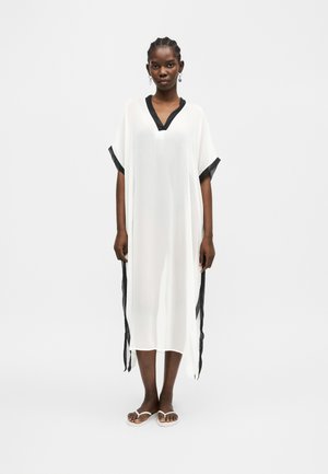 Naine seisab, kandes lahtist valget kleiti musta äärise ja valgete sandaalidega, heleda tausta ees.