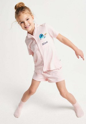 Junges Mädchen lächelt, trägt hellrosa Stitch-Pyjamas mit kurzen Ärmeln und Shorts, steht mit gespreizten Beinen und ausgestreckten Armen.