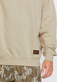 Sudadera beige hecha de una tela suave con puños acanalados. Presenta una etiqueta negra y naranja en el dobladillo. Se lleva con pantalones de camuflaje marrón con estampado.