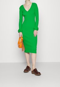 Grünes Bodycon-Kleid mit V-Ausschnitt und langen Ärmeln, die mit Rüschensäumen versehen sind, kombiniert mit einer strukturierten orangefarbenen Handtasche und braunen Clogs.