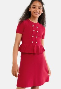 Rød strikket kjole med peplum-talje, korte ærmer, knapdetaljer og en struktureret overflade. Accentueret med dekorative knapper.