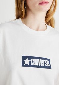 Weißes Baumwoll-T-Shirt mit einem navyblauen rechteckigen Logo, das einen Stern und das Wort "Converse" in fetten Buchstaben zeigt. Einfacher Rundhalsausschnitt.