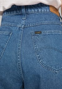 Jeans in denim blu con vita alta, caratterizzati da una patch in pelle del marchio Lee, tasche posteriori e dettagli di cucitura visibili.
