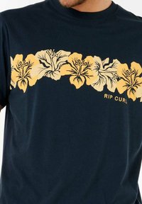 Navyblaue Baumwoll-T-Shirt mit einem gelben Hibiskusblüten-Graphic über der Brust. "RIP CURL"-Logo in Gelb unten rechts.