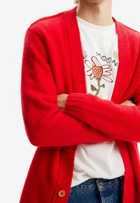 Cardigan rosso in lana con una texture morbida, caratterizzato da una vestibilità lunga, polsini a coste e bottoni in legno. Indossato sopra una camicia bianca con ricami floreali.