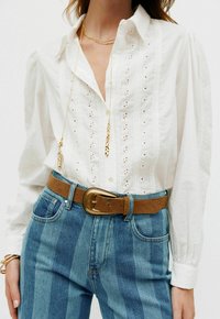 Hvid knap-up skjorte med hulmønster-detailing, lange ærmer, båret med blåstribede jeans og et brunt bælte med en stor spænde.