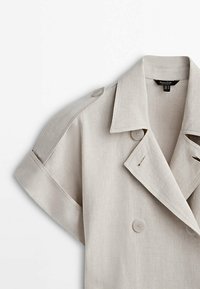 Jasno beżowa lniana kurtka z podwójnym zapięciem, krótkimi rękawami i naramiennikami. Posiada akcenty guzików oraz standardowy kołnierz.