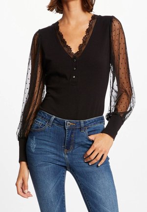 Frau trägt ein schwarzes Oberteil mit transparenten gepunkteten Puffärmeln und Spitzenbesatz, kombiniert mit blauen, hoch taillierten Jeans mit leichter Used-Optik.