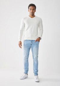 Witte v-hals trui, lichtblauwe spijkerbroek en witte sneakers. De outfit heeft een aangesloten pasvorm en een gladde, zachte textuur.