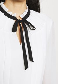 Blouse blanche avec col plissé et nœud papillon noir au niveau du décolleté sur une personne aux cheveux longs et foncés.