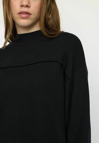 Pulover negru cu guler înalt, textură cu nervuri și o cusătură orizontală pe piept, cu umere căzute și croială lejeră.
