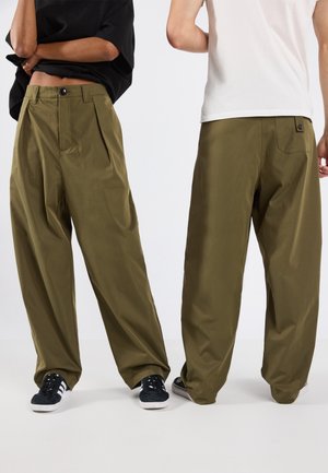 Twee personen met losse olijfgroene broeken en sneakers, één naar voren gericht met een zwart shirt, de ander met een witte blouse naar achteren gekeerd.