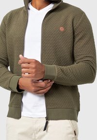 Sweat à capuche zippé vert olive avec design texturé matelassé, poignets côtelés et écusson logo sur la poitrine, porté sur un T-shirt blanc.