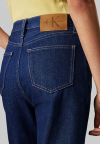 Jeans de mezclilla de un azul oscuro con una textura suave, que cuentan con una etiqueta de cuero de Calvin Klein en la cinturilla y bolsillos traseros estándar.