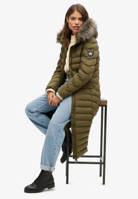 Olijfgroene gewatteerde jas met een met bont gevoerde capuchon, lange lengte, ritssluiting en logo-embleem, gecombineerd met lichtblauwe jeans en zwarte enkellaarsjes.