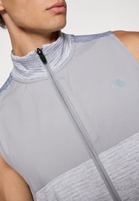 Gray sleeveless jacket with a high collar, featuring a smooth texture, contrasting lighter gray stripes, and a zip closure. New Balance logo present.