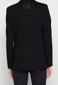 Černý blejzr se strukturovaným designem, s hladkým povrchem, klopami do tvaru V a výraznou logovkou na zadní straně. Úzký střih, manžety na knoflíky.