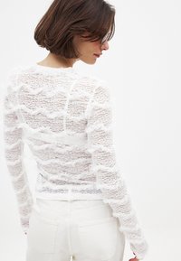 Top bianco in pizzo con un motivo ondulato e dettagli frangiati, con maniche lunghe. Abbinato a pantaloni in denim bianchi. Design semplice e strutturato.