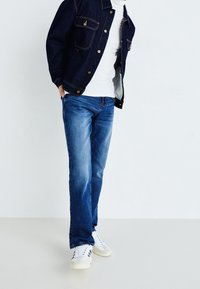 Mörk denimjacka med guldstygn, vit T-shirt och blå jeans, kombinerat med vita sneakers med svarta detaljer.