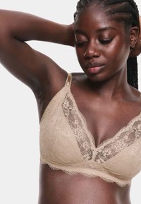 Bralette de encaje en beige claro, con diseño de escote en V, tirantes ajustables, patrón floral y bordes en forma de ondas, con una textura suave.