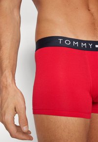 Rote Baumwoll-Boxershorts mit einem schwarzen elastischen Bund, der das "TOMMY"-Logo in Weiß trägt. Glatte Textur, körpernahe Passform und Länge bis zur Mitte des Oberschenkels.