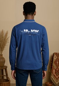 Blauwe langesleeve shirt met een klassieke kraag, voorzien van witte geprinte graphics op de achterkant en met knoopsluiting bij de manchetten. Zachte katoenen textuur.
