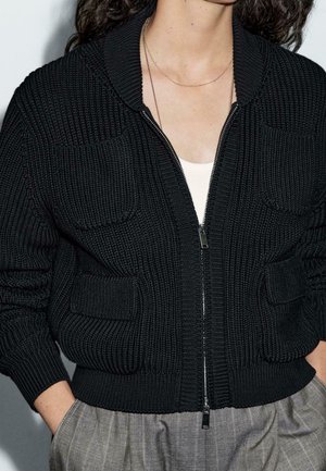 Femme portant une veste en maille noire à fermeture éclair avec poches avant, par-dessus un haut blanc et un pantalon à carreaux gris, mains dans les poches.