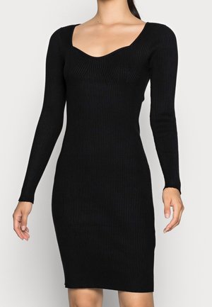 Woman wearing a fitted black ribbed knit dress with long sleeves and a square neckline, shown from neck to knees.