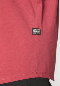 T-shirt en coton rouge avec un ourlet arrondi, arborant une étiquette noire G-Star Originals cousue sur le côté pour l'identification de la marque.