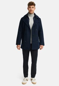 Marineblå peacoat med dobbeltknapning og store kraver, båret over en lys grå turtleneck og parret med mørke bukser og sneakers.