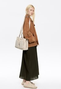 Sac à main beige avec deux poches avant, associé à une veste en daim marron, une jupe vert foncé et des chaussures de couleur claire. Textures lisses et tons neutres.