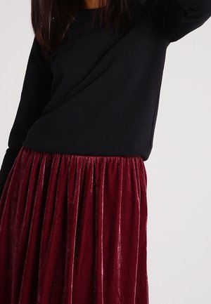 Woman wearing a long-sleeve black top and a gathered, shiny deep red velvet skirt against a plain light background.
