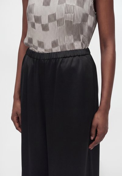 Femme portant un top sans manches à motifs, rentré dans un pantalon noir ample taille haute avec une ceinture élastique.