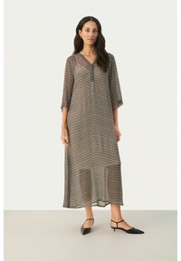 Robe longue et translucide avec un motif en zigzag noir et beige. Elle présente un décolleté en V et des manches trois-quarts. Portée avec des chaussures noires à talons pointus.