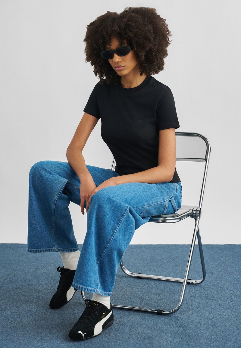 Woman with curly hair wearing black sunglasses, black t-shirt, blue wide-leg jeans, and black sneakers sitting on a metal folding chair.