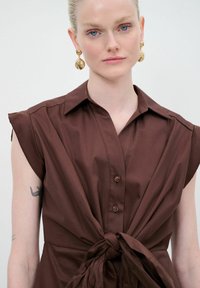 Abito marrone a maniche corte con dettaglio in vita annodato, dotato di colletto a camicia e bottoni marroni, realizzato in un tessuto liscio.