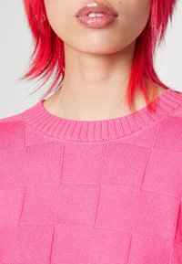 Klart rosa stickad tröja med ett strukturerat rutmönster, ribbad halskrage och slät yta. Lämplig för vardagsbruk.