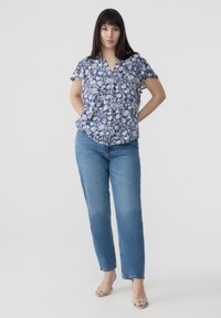 Blusa floreale blu navy con motivi bianchi, maniche corte a volant, scollatura a V, abbinata a jeans dritti di media tonalità e sandali verde chiaro.