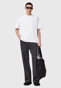 Camiseta blanca de manga corta, pantalones vaqueros negros holgados, zapatos negros tipo slip-on y una bolsa negra con correas para el hombro. Elementos de diseño minimalista.