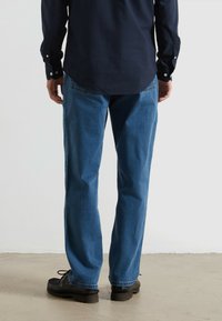 Jean en denim bleu avec une coupe droite, tissu délavé et design à cinq poches. Associé à une chemise à boutons sombre et des chaussures noires.