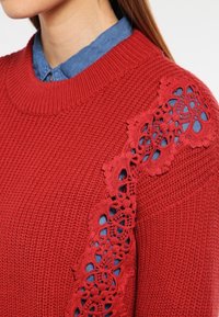 Maglione rosso a maglia con corpo texturizzato e dettagli in pizzo intricati sulla spalla, indossato sopra una camicia in chambray blu.